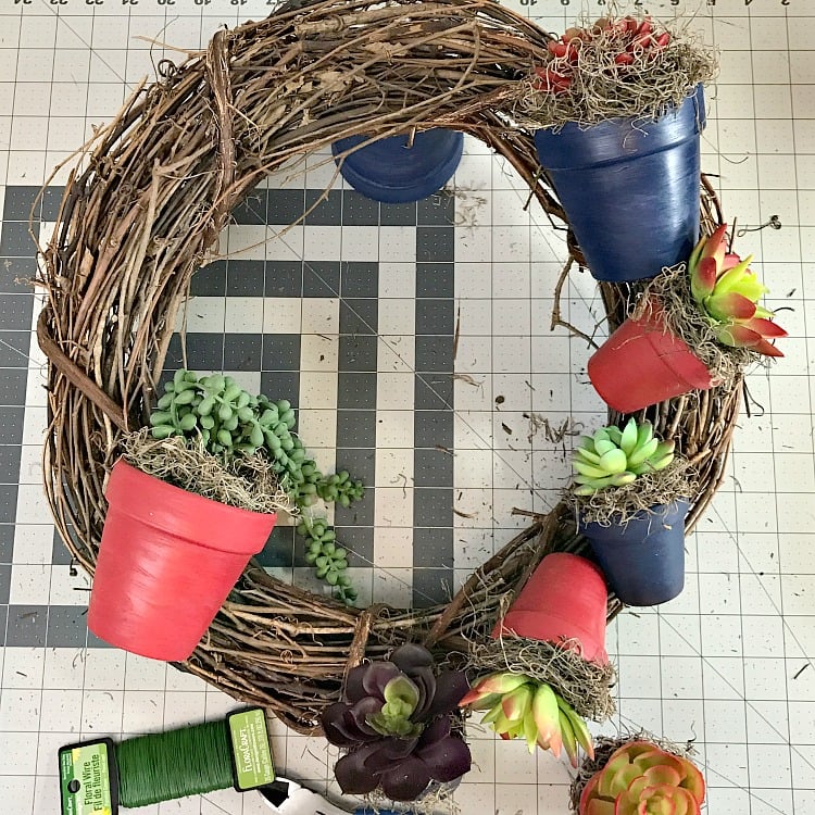 A grapevine wreath with succulent filled painted clay flower pots being affixed to it.
