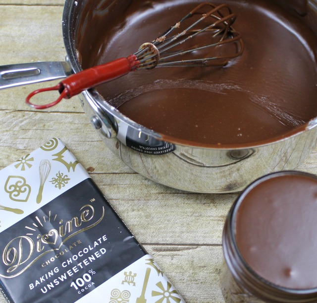 Photo of a sauce pan filled with fresh Homemade Chocolate Fudge sauce next to a bar of baking chocolate and a mason jar filled with the sauce all sitting on a wooden surface