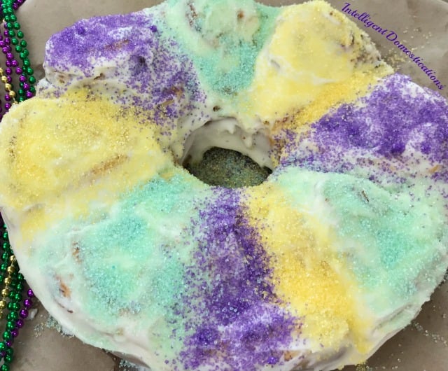 King Cake with purple, yellow and green sugar crystals on top of heavy icing with Mardi Gras beads next to it