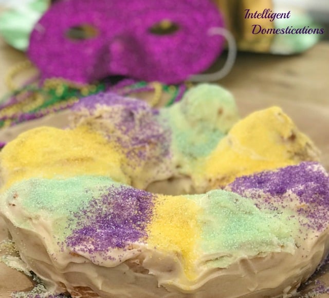 A King Cake made with cinnamon rolls and covered in purple, yellow and green sugar sprinkles with a Mardi Gras mask in the background