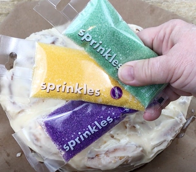 Purple, yellow and green sugar sprinkles held over a King Cake
