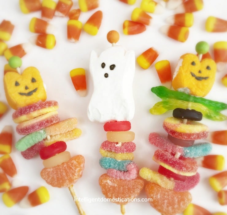 Make these grab and go Halloween Party Treats! Our Candy Kabobs are easy to make with wooden candy sticks, marshmallow ghosts and gummy candies! Halloween candy kabobs displayed on a white background surrounded by candy corn.