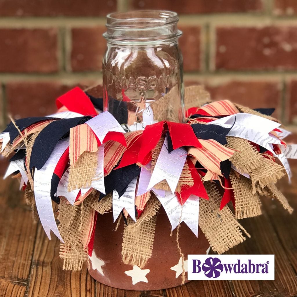 A red white and blue centerpiece which is a terracotta clay pot with white stars on the bottom, ribbons spilling out and a Mason jar candle in the center. Pictured outside on a wood table with a brick wall in the background