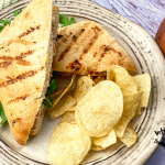 Panini cut in half with potato chips on a white plate and a jar of Peach sauce on a wooden table
