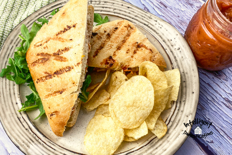 Panini cut in half with potato chips on a white plate and a jar of Peach sauce on a wooden table