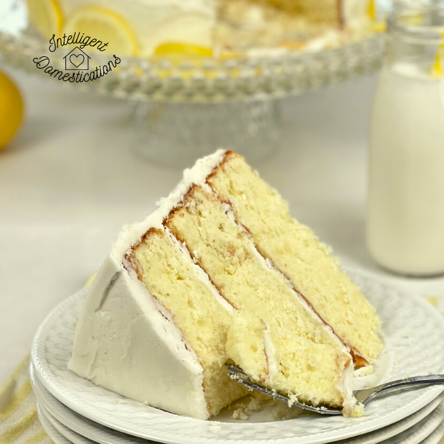 a piece of three layer lemon cake on a plate in front of the cake stand and a bottle of milk